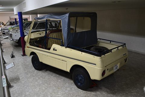 Rear quarter of a cream small utility with a navy canvas top over the seats and a spare wheel in its load bed, on a museum floor. Opens a larger view