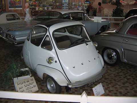 Off-white bubble car among larger classic cars on a tiled museum floor, a handwritten do-not-touch sign by its wheel.
