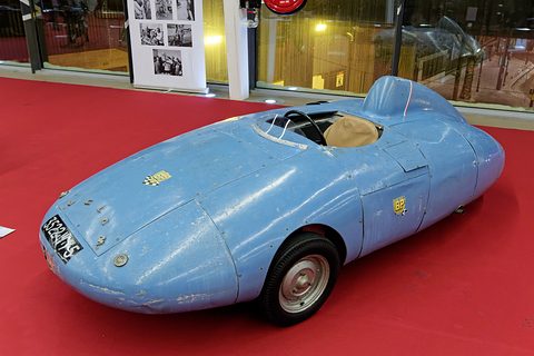 Worn pale blue aluminium streamliner with a tiny windscreen, tan seat and head fairing, seen from the front left on red carpet. Opens a larger view