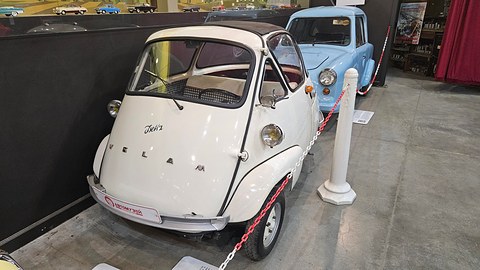 White egg-shaped car with Isetta script and spaced letters on its door, behind a red and white chain, a pale blue car beside it.