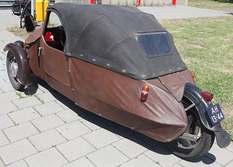 Rear quarter of a brown three-wheeler: black hood with rear window, single rear wheel and blue number plate. Opens a larger view