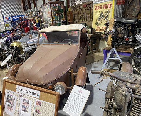 Brown leatherette three-wheeler with beige hood among motorcycles and signs in a crowded museum shed. Opens a larger view