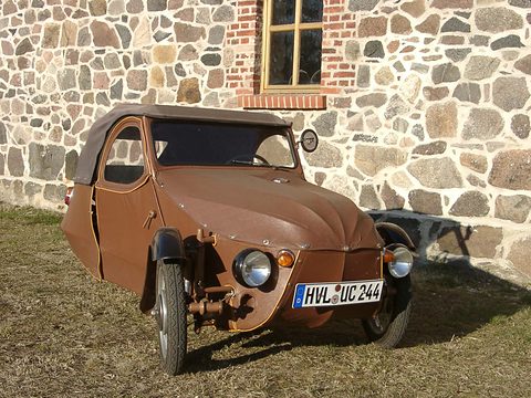 Brown leatherette three-wheeler with exposed front wheels and round headlamps, parked on grass before a fieldstone wall. Opens a larger view