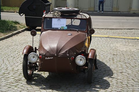 Front of a brown leatherette three-wheeler with twin headlamps, Velorex script and spare wheel on top, on cobbles. Opens a larger view