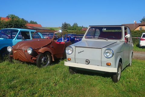 Brown open three-wheeler with roof hoops next to a boxy grey microcar with white bumper, on grass. Opens a larger view
