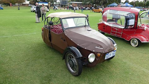 Brown three-wheeler with beige hood, red pleated seats and spoked wheel, beside a red bubble car on grass. Opens a larger view