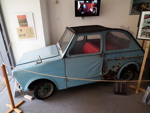 Faded pale blue two-door microcar with black fabric roof and red seat, behind a rope in a white museum room. Opens a larger view