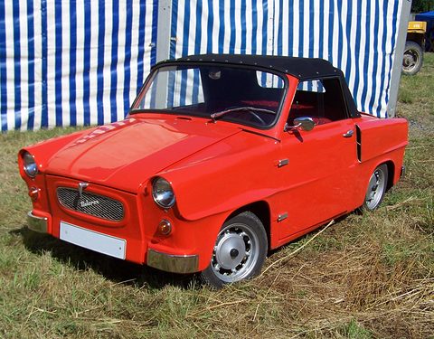 Small red open two-seater with black hood, oval grille with Velorex script, parked on grass before a striped tent. Opens a larger view