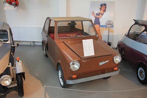 Boxy brown two-door microcar with beige roof, round headlamps and Velorex badge, between other small cars.