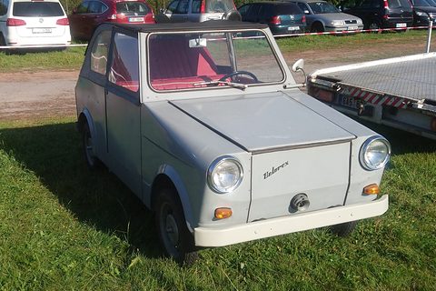 Grey boxy two-door microcar with dark roof, red interior and round headlamps, parked on grass by a trailer. Opens a larger view