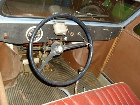 Thin black steering wheel before a black fascia with one round speedometer, ribbed rubber floor and red bench. Opens a larger view