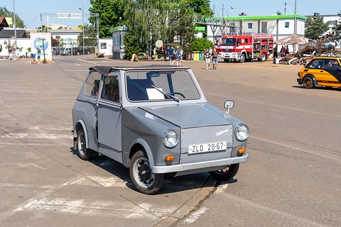 Grey boxy two-door microcar with black fabric roof and white front plate on a sunny concrete yard. Opens a larger view
