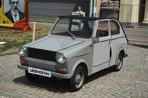 Boxy grey two-door microcar with black fabric roof, round headlamps and event plate, on cobbles beside a lawn. Opens a larger view