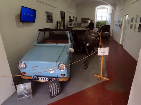 Front of a faded pale blue microcar with Velorex script and French plate, information boards and other exhibits behind. Opens a larger view