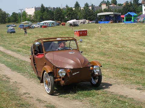 Brown three-wheeler with roof hoops and Velorex script driving along a dirt track on a grass field. Opens a larger view