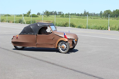 Brown three-wheeler with black hood and roof rack moving across grey tarmac, green field and fence behind. Opens a larger view