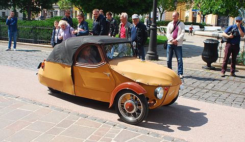 Ochre leatherette three-wheeler with black hood and red wings driving on paving past watching spectators. Opens a larger view