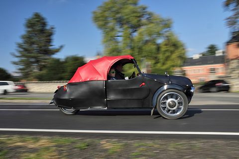 Black three-wheeler with red rear hood driving fast along a road, panned so the trees behind are blurred. Opens a larger view