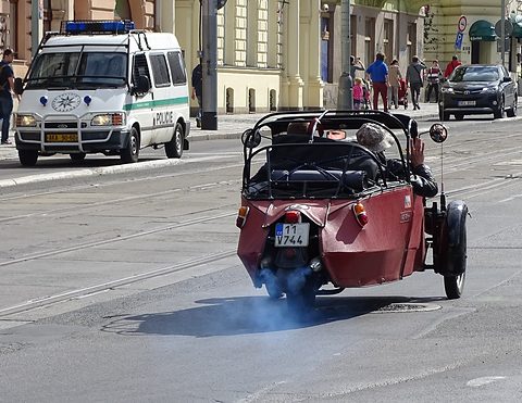 Red open three-wheeler with roof hoops driving away on a city street, blue exhaust smoke behind it. Opens a larger view
