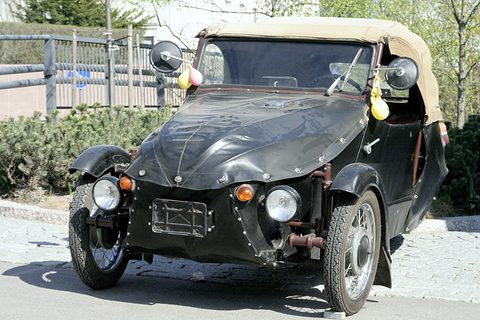 Black leatherette three-wheeler with beige hood, round mirrors with balloons tied on, spoked wheels, by a hedge. Opens a larger view
