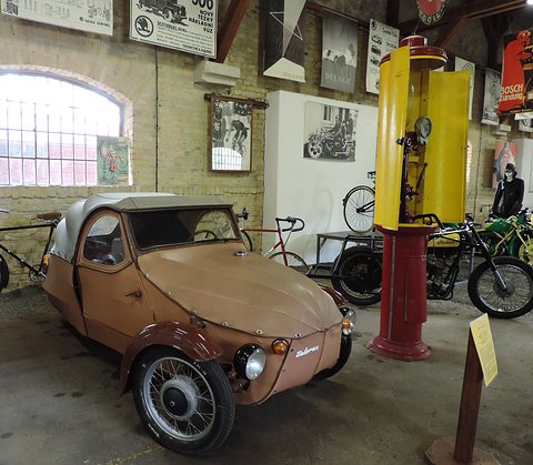Tan fabric three-wheeler with beige hood and brown wings in a brick hall, yellow petrol pump and bicycles beside it. Opens a larger view