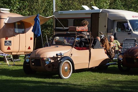 Tan three-wheeler with roof hoops, suitcase and leather bag on the back, parked on grass by a caravan and campers. Opens a larger view