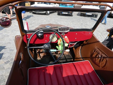 View over a red bench seat to a red dashboard with large black steering wheel and round speedometer. Opens a larger view