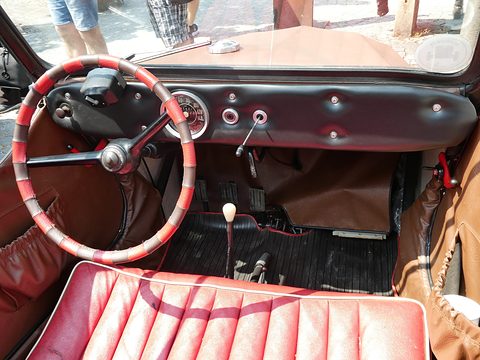 Padded black dashboard with round speedometer, red-bound steering wheel, floor gear lever and red bench seat. Opens a larger view