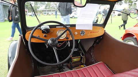 Wooden fascia with speedometer, round gauge, black steering wheel, exposed battery and red pleated bench. Opens a larger view