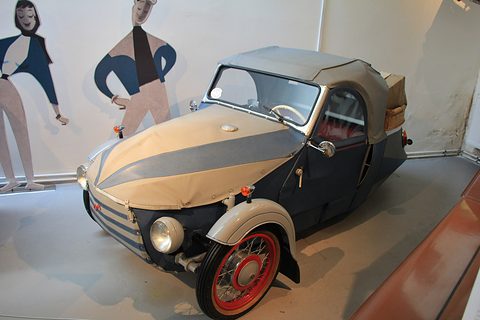 Striped cream and blue-grey fabric three-wheeler with hood up and basket on the back, cartoon figures on the wall. Opens a larger view
