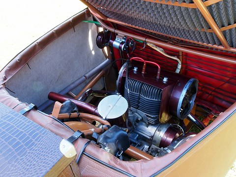 Two-cylinder motorcycle engine with maroon fan housing inside an orange tube frame, padded cover lifted above. Opens a larger view