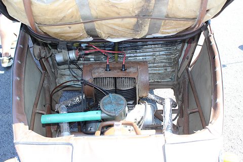Rusty two-cylinder engine with green air intake, wiring and exhaust pipes inside a brown steel-tube frame. Opens a larger view