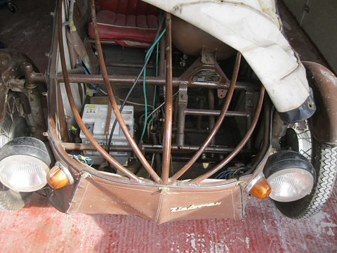 Brown painted tubes curving to the nose under a lifted white cover, with battery, wiring and front headlamps. Opens a larger view