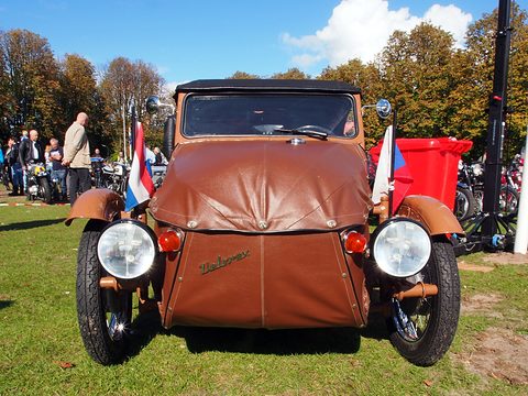 Front of a brown leatherette three-wheeler with black hood, twin headlamps, Velorex script and two small flags. Opens a larger view