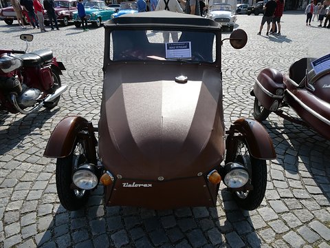 Front of a dark brown three-wheeler with black hood, twin headlamps and Velorex script on cobbles. Opens a larger view