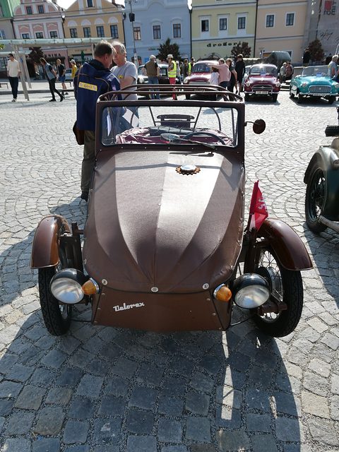 Front of a brown three-wheeler with twin headlamps, metal wings and Velorex script, on a cobbled square with people behind. Opens a larger view