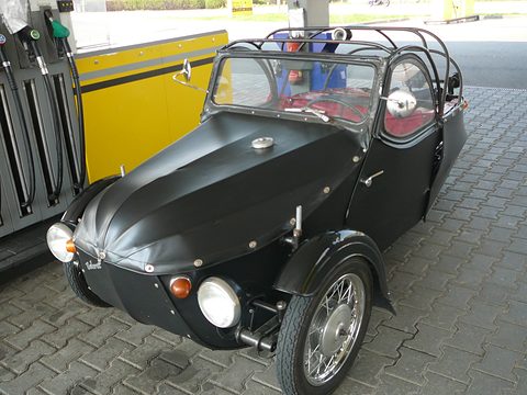 Black fabric-bodied three-wheeler with roof hoops, chrome headlamps and spoked wheel beside yellow fuel pumps. Opens a larger view
