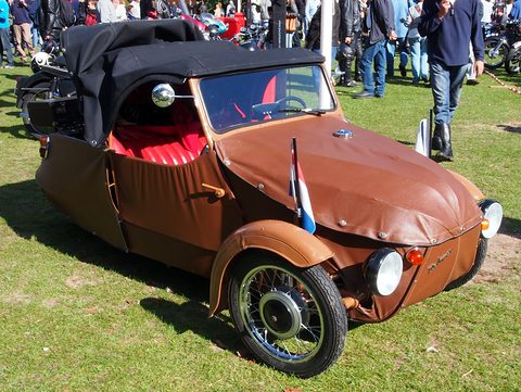 Brown fabric-bodied three-wheeler with black hood, red seats, spoked wheel and small flag on the wing, on grass. Opens a larger view