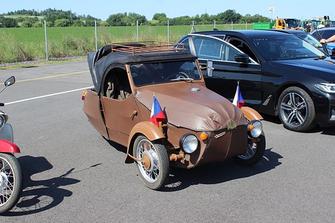 Brown leatherette three-wheeler with black hood, roof rack and two small flags beside a black modern saloon. Opens a larger view