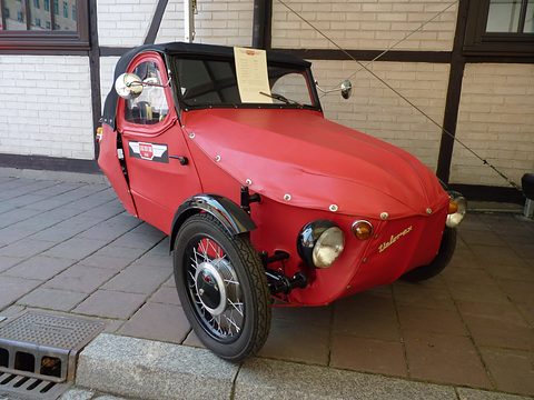 Red leatherette three-wheeler with black fabric hood, spoked front wheel and info sheet in the windscreen, by a half-timbered wall. Opens a larger view