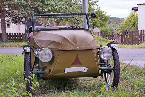 Olive leatherette three-wheeler with green frame, chrome headlamps and a brown triangle on the nose, on grass. Opens a larger view