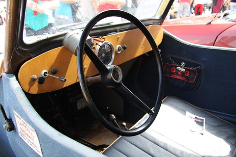 Wooden fascia with one round speedometer, large black steering wheel, floor gear levers and pale bench seat. Opens a larger view