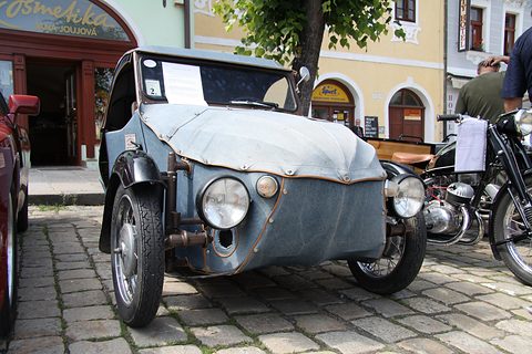 Front of a blue-grey fabric three-wheeler with twin headlamps and exhibition sheet in the windscreen, beside a motorcycle. Opens a larger view