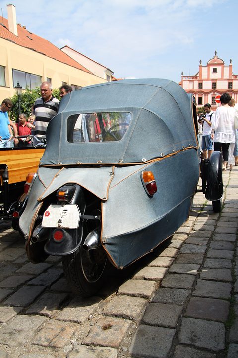 Rear of a blue-grey fabric-bodied three-wheeler on cobbles, rear window in the hood, visitors around it. Opens a larger view