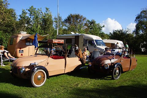 Two open three-wheelers with roof hoops and luggage on grass, a small tan caravan and campers behind. Opens a larger view