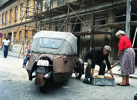 Rear view of a small fabric-bodied three-wheeler by scaffolding, an elderly man lifting a crate and a woman beside him. Opens a larger view