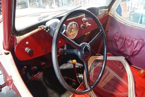 Black three-spoke steering wheel, cream speedometer on a maroon dashboard, red bench seat and a loose side window. Opens a larger view