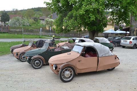 Row of tan, green and brown three-wheelers parked on gravel beside a village road under a big tree. Opens a larger view