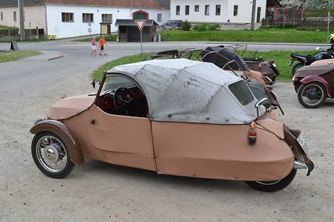 Tan three-wheeler with grey fabric hood seen from the rear quarter, other Velorexes parked along a village road. Opens a larger view