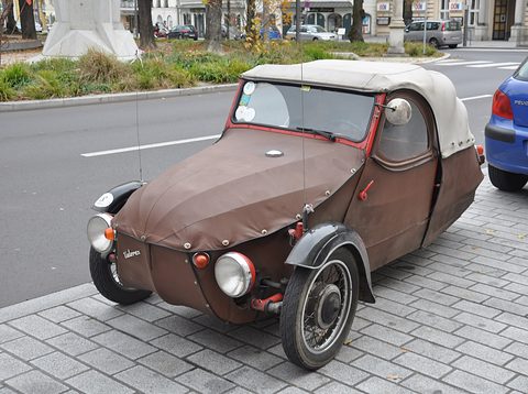 Brown fabric-bodied three-wheeler with grey hood, Velorex script and spoked wheel on paving beside a blue hatchback. Opens a larger view